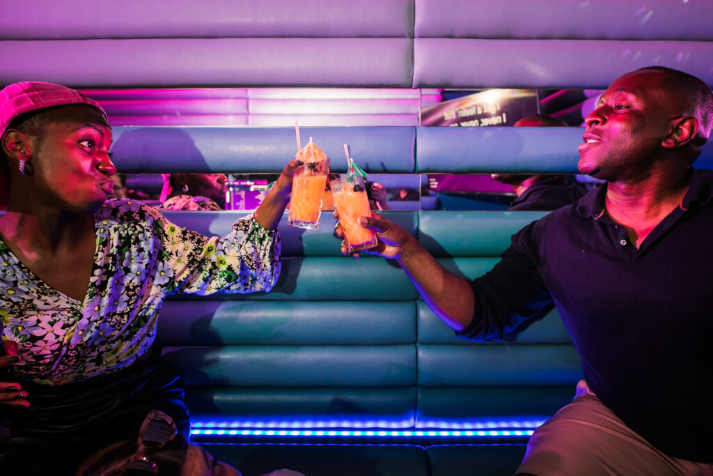 Two friends clinking tropical cocktails inside a neon-lit, private karaoke pod at Lucky Voice Waterloo.