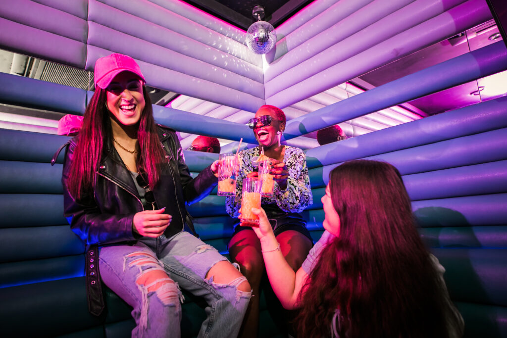 Three young women in a small, private karaoke room, complete with a disco ball, holding cocktails and laughing, surrounded by brightly lit pink and blue ribbed walls.
