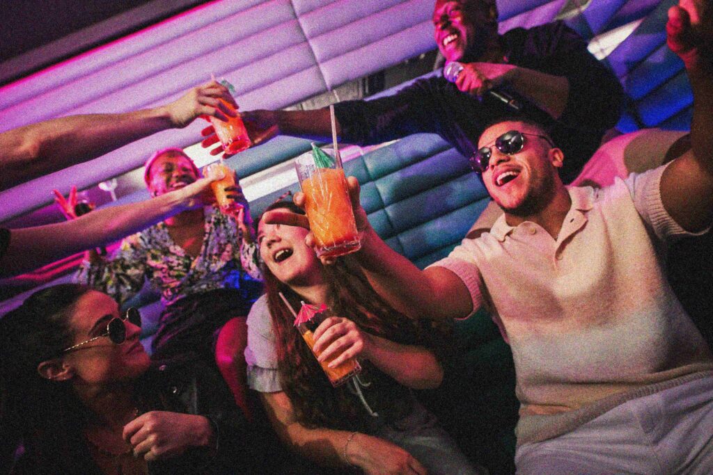 A group of friends cheering, singing into a microphone, and raising cocktail glasses inside a vibrant, pink-and-blue lit private karaoke room at Lucky Voice Waterloo.