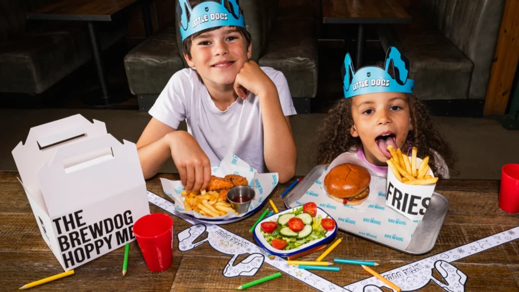 Two children wearing paper crowns enjoying kids' meals at a BrewDog dining table.