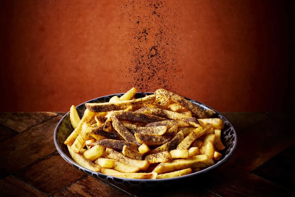 A bowl of golden-brown Nando’s chips being sprinkled mid-air with dark, smoky black seasoning, resting on a rustic wooden table against a warm orange-brown background.