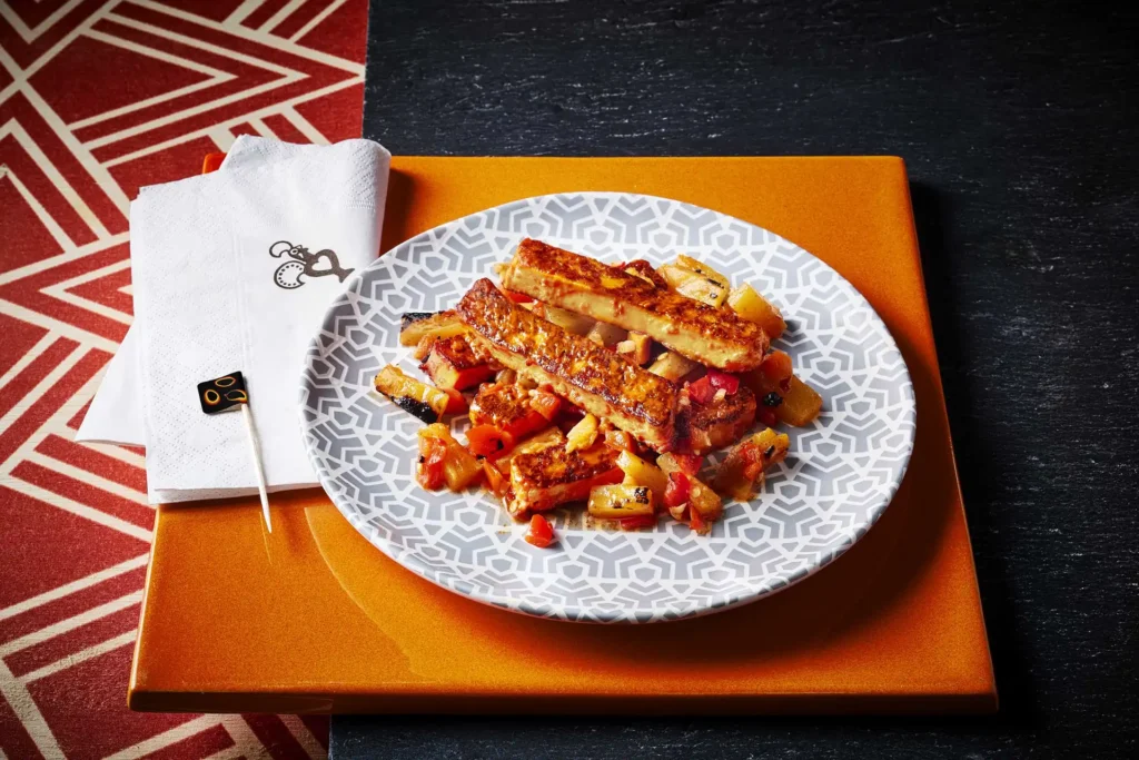 Grilled Nando’s halloumi sticks drizzled in red sauce and served over diced salsa on a patterned plate, resting on a bright orange square surface next to white napkins and a small black cocktail pick featuring orange flame icons.