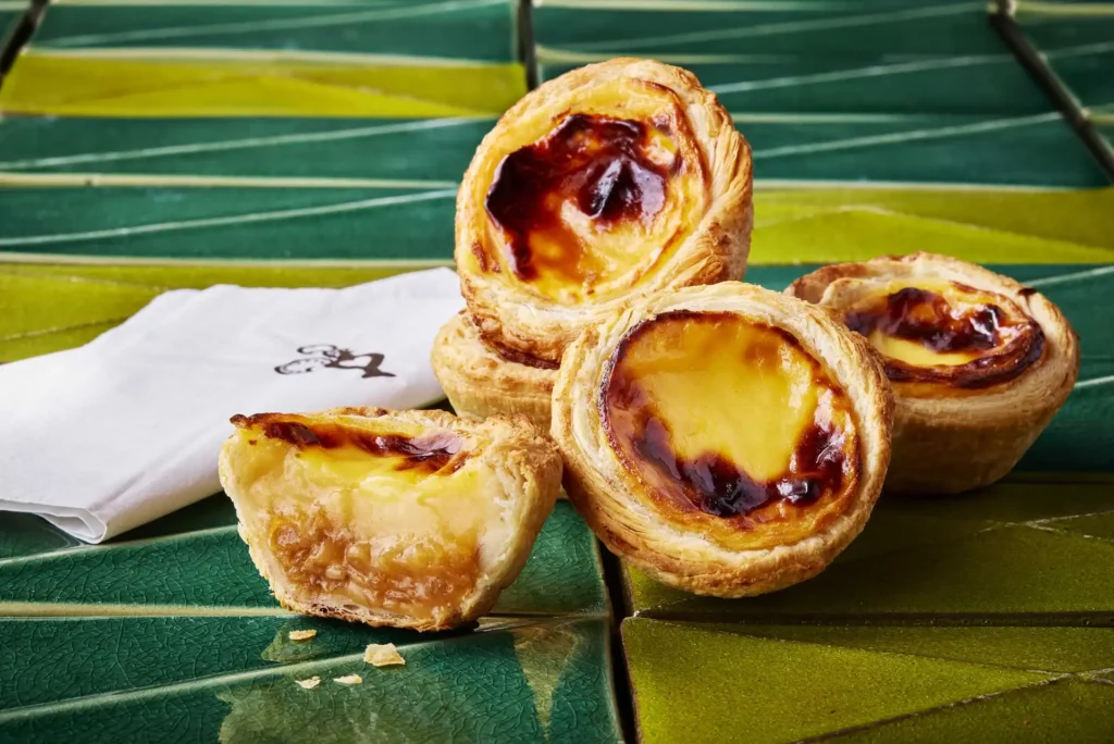 A stack of golden, flaky Nando’s Toffee Apple Pastel de Natas with caramelized, blistered custard tops. One tart is cut in half in the foreground, showing a sweet apple and toffee filling beneath the custard layer, set against a green tiled backdrop next to a white napkin.